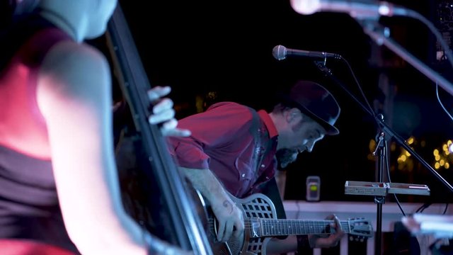 Looking through the female on the double bass, a male roots musician rocks out on a resonator guitar at night at a restaurant.