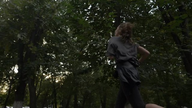 Beautiful girl with curled long hair running near the trees in park to loose extra weight in slow motion