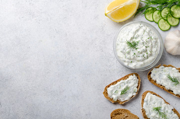 Homemade greek tzatziki sauce in a glass bowl with ingredients and sliced bread on a light stone background. Cucumber, lemon, dill, garlic. Top view, horizontal, copy space