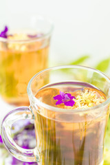 Linden herbal tea in a transparent grog glass with a linden blossom and bunch of herbs on the white wooden surface