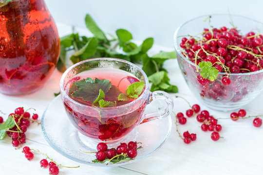Redcurrant drink in transparent glass carafe and cup. Clear glass vase with red currant berries on the white wooden background.