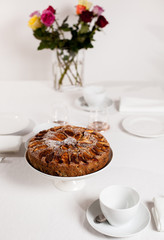 Fresh homemade apple cake on a laid table with white table cloth and a vase of roses, shot from above