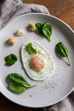 Single fried egg in egg shape over white plate garnished with spinach, green herbs, salt, papper, horseradish and hummus over wooden bord with grey napking on the side