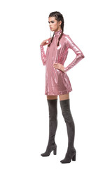 .Young lady looking aside while standing in pink dress in sequins and knee high boots on white background.