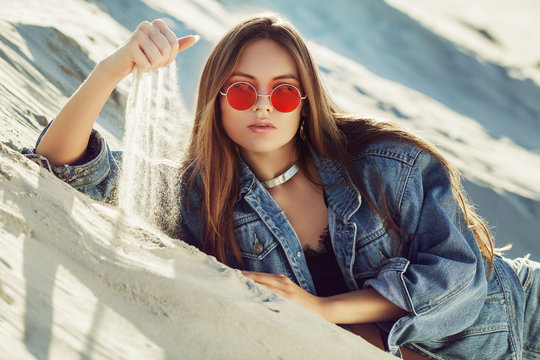 Portrait of young beautiful woman wearing stylish light blue denim jacket, red round sunglasses. Model posing and playing with sand. Female fashion conept 