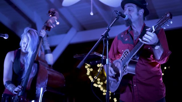 Resonator guitarist plays and sings into a microphone while a female bass player accompanies at an outdoor cafe performance at night.