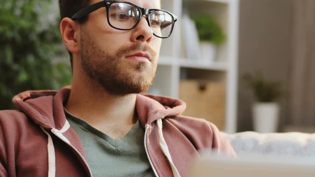 Close up of the handsome young man with beard in glasses thinking about something and orking on the laptop in the living room. Indoors. Portrait