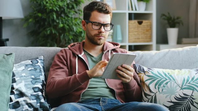 Attractive young man in glasses scrolling and taping on his tablet device while sitting on the sofa in the nice living room. Working at home. Indoor