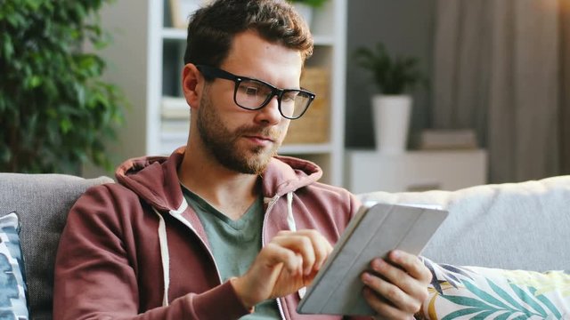 Portrait shot of the young stylish man with beard holding a tablet computer and scrolling on it in the modern nice room at home. Indoors