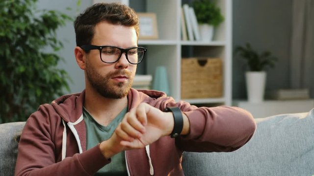 Portrait shot of the young handsome caucasian man rising his hand with a smart watch on and taping on its screen while sitting on the sofa in the nice room at home.. Indoor