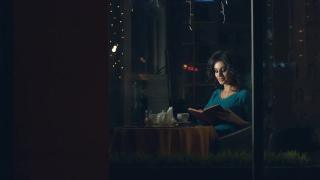 Cute womanin in a cafe.
A pretty woman is sitting in a cafe behind the glass and reading a book.
The glass reflects the traffic of urban transport.