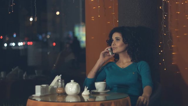 Girl with a smartphone in a cafe.
Slow motion. A pretty girl is sitting in a cafe and talking on the phone. She smiles.
Outside the window lights the evening city.