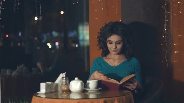 Girl with a book in a cafe.
Slow motion. A pretty girl is sitting in a cafe and reading a book.
Outside the window lights the evening city.