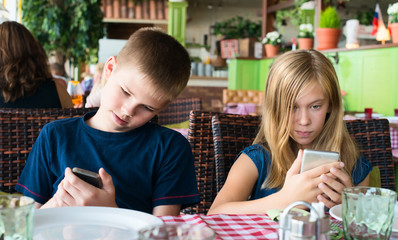 Teenagers having fun with mobile phones in cafe. Modern lifestyle and technology concept. Children sitting in restaurant and looking on the smartphones.