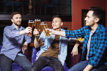 Three young men in casual clothes are smiling and clanging glasses of beer together while sitting in pub
