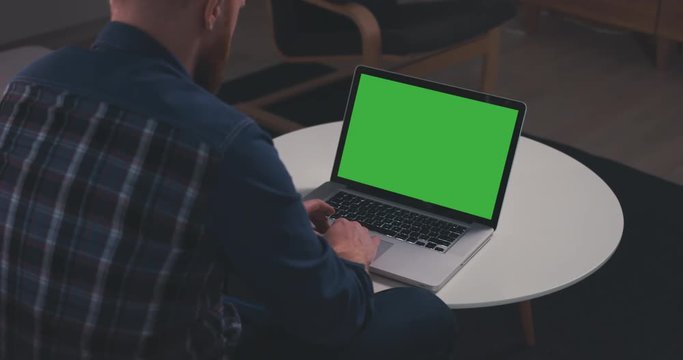 Over the shoulder shot of a young Caucasian man sitting on sofa, working from home on a laptop, green screen chroma key. 4K UHD 60 FPS SLO MO