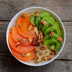Oatmeal porridge with fresh sweet persimmon, kiwi fruit and raw walnuts in a round white bowl...