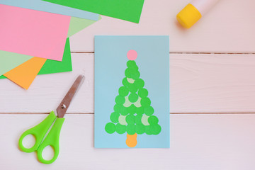 Paper Christmas greeting card made of simple geometric shapes, colored paper set, scissors, glue stick on a wooden table. How to make beautiful Christmas card. Children creative development. Top view