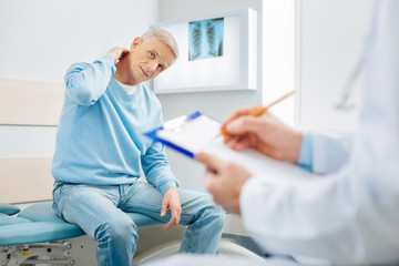 Medical conclusion. Nice optimistic senior man looking at his therapist and touching his neck while...