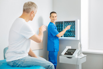 Fototapeta premium Diagnosis explanation. Cheerful delighted nice doctor standing near the X ray images and pointing at them while talking to his patient