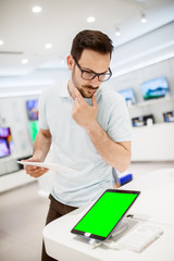 Curious young bearded handsome man looking and thinking about the new tablet in a tech store with...