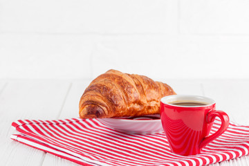 Freshly baked croissant on napkin, cup of coffee in red cup on white wooden background. French breakfast. Fresh pastries for breakfast. Delicious dessert. Closeup photography. Horizontal banner