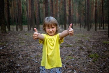 Little girl with thumbs up in the pine forest in summer.