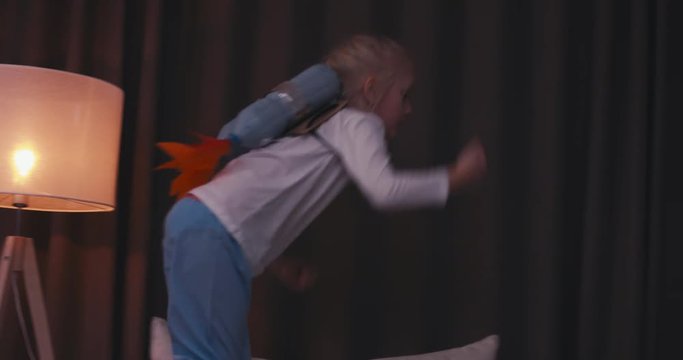 Funny cute dreamer girl wearing rocket pack made from plastic bottles pretending to be an astronaut or pilot, jumping from sofa. 4K UHD 60 FPS SLO MO