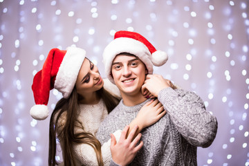 Cheerful attractive couple in winter clothes and santa hats hugging and kissing on bokeh light background