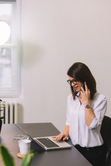 Portrait of pretty young woman working with laptop in her office. Pretty girl talking on her mobile in her office.