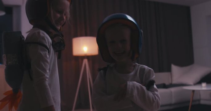 Funny cute dreamer siblings wearing helmets and rocket packs made from plastic bottles pretending to be astronauts, jumping from sofa. 4K UHD 60 FPS SLO MO