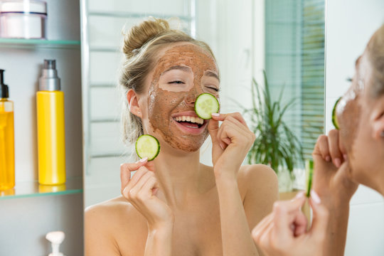 Young beautiful girl applying facial scrub mask  and slice of cucumber on skin. Looking in the mirror in bathroom, Wrapped in a towel, having fun.
