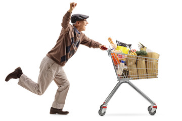 Joyful elderly man pushing a shopping cart filled with groceries and holding his hand up