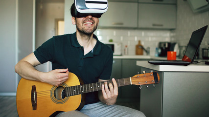 Happy young man sitting at kitchen learning to play guitar using VR 360 headset and feels him guitarist at concert at home