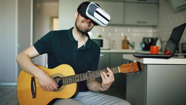 Happy young man sitting at kitchen learning to play guitar using VR 360 headset and feels him guitarist at concert at home