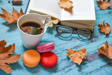 Hot coffee, glasses, macaroons and book with autumn leaves on blue wood background - seasonal relax concept. Flat lay