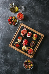 Tapas or bruschetta variety. Crusty bread with ham prosciutto, sun dried tomatoes, olive oil, olives, pepper on slate wooden serving board over dark texture background. Top view with space