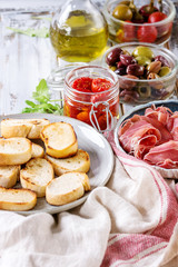 Ingredients for making tapas or bruschetta. Crusty bread, ham prosciutto, sun dried tomatoes, olive oil, olives, pepper, greens on plates over white plank wooden table. Close up
