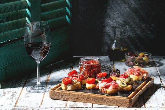 Fototapeta Tapas or bruschetta variety. Bread with ham prosciutto, sun dried tomatoes, olive oil, olives, pepper on slate wood board with glass of red wine over white table. Light via old jalousie. Rustic style.