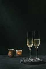 Two glasses of white champagne served on black wooden decorative board with candles over dark texture background. Copy space