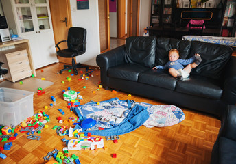 Adorable 1 year old baby boy with funny facial expression playing in a very messy living room, lying on a couch, watching tv