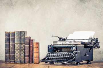 Vintage old books and aged black typewriter with paper blank on wooden table front concrete wall...