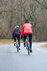 Fototapeta na wymiar Two Young Female Cyclists Riding Road Bicycles in the Park in the Cold Autumn Morning. Healthy Lifestyle.