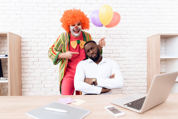 A man in a clown suit is standing beside a black man who sits at his desk.