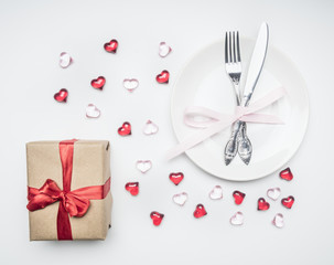 composition by February 14, white dish with knife and fork, bow, gift and hearts laid out around, on white background, top view