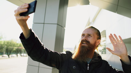 Young bearded hipster man having online video chat with smartphone camera while travel city street
