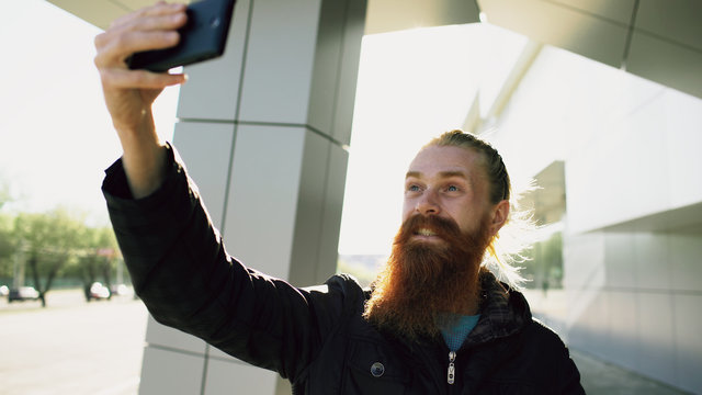 Young bearded hipster man taking selfie picture using smartphone camera outdoors at city street