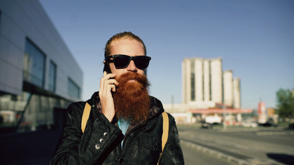 young bearded hipster man in sunglasses smiling and talking smartphone while travelling city street