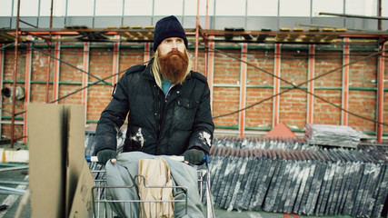 bearded homeless man walking down the street with shooping card at cold autumn day