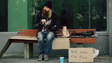 Homeless and jobless european man with cardboard sign eat sandwich on bench at city street because of immigrants crisis in Europe